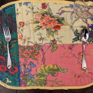 Beautiful handcrafted patchwork placemat with vibrant floral patterns in red, pink, yellow, and green. The placemat is finished with a yellow trim, adding a cheerful touch to any table setting. Perfect for enhancing your dining decor. Made in the USA Handcrafted multicolor cotton placemat set with vibrant floral panels and yellow edging, styled on a reclaimed Tropical floral print placemat close‑up in quilted cotton patchwork, ideal for summer dinner parties, and everyday use wood dining table
