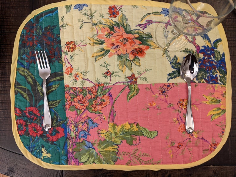Beautiful handcrafted patchwork placemat with vibrant floral patterns in red, pink, yellow, and green. The placemat is finished with a yellow trim, adding a cheerful touch to any table setting. Perfect for enhancing your dining decor. Made in the USA Handcrafted multicolor cotton placemat set with vibrant floral panels and yellow edging, styled on a reclaimed Tropical floral print placemat close‑up in quilted cotton patchwork, ideal for summer dinner parties, and everyday use wood dining table