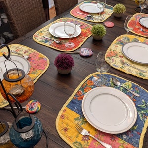 Crafted in the USA, this reversible placemat set showcases a bright and colorful patchwork design with floral patterns in red, yellow, blue, and pink. Ideal for bringing a festive and vibrant touch to your dining decor. Quilted cotton placemats set featuring vibrant garden‑inspired panels in coral pink, emerald green, navy blue, and buttercup yellow