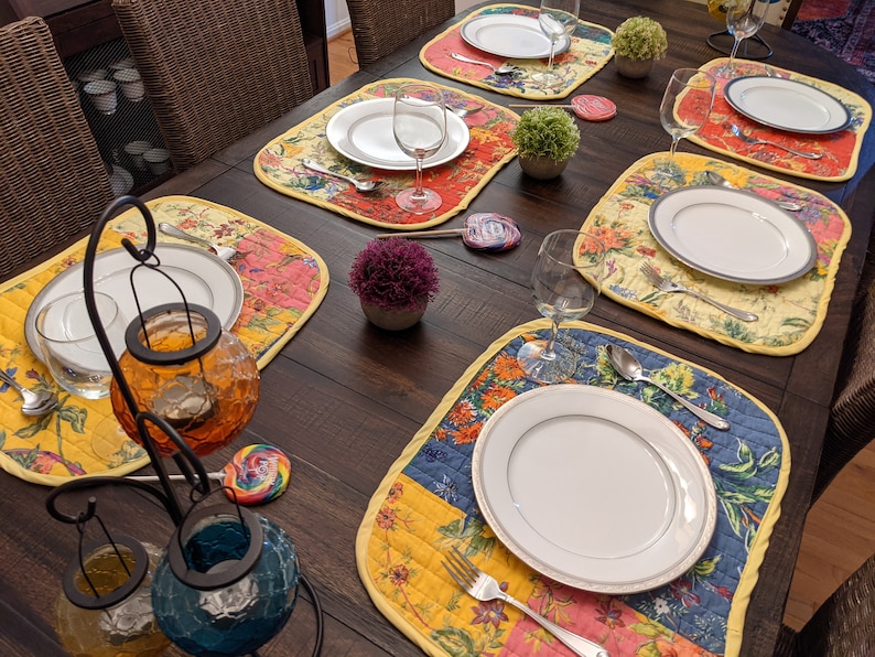 Crafted in the USA, this reversible placemat set showcases a bright and colorful patchwork design with floral patterns in red, yellow, blue, and pink. Ideal for bringing a festive and vibrant touch to your dining decor. Quilted cotton placemats set featuring vibrant garden‑inspired panels in coral pink, emerald green, navy blue, and buttercup yellow