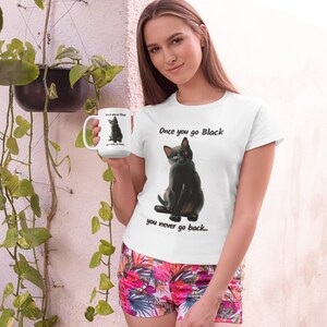 May include: White t-shirt with a black cat graphic and the text "Once you go Black you never go back...". The image also shows a white mug with the same cat graphic. The person is wearing floral shorts.