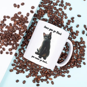 May include: White ceramic mug featuring a black cat illustration with the text "Once you go Black you never go back..." surrounded by coffee beans. The mug has a white handle and is set against a light blue and white background.