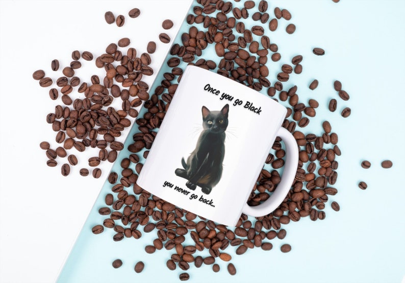 May include: White ceramic mug featuring a black cat illustration with the text "Once you go Black you never go back..." surrounded by coffee beans. The mug has a white handle and is set against a light blue and white background.