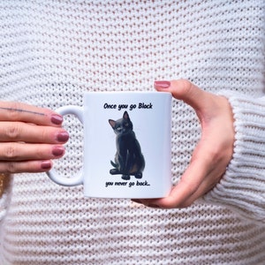 May include: White ceramic mug featuring a black cat illustration with the text "Once you go Black, you never go back...". The mug has a curved handle and is held by a person wearing a white sweater.