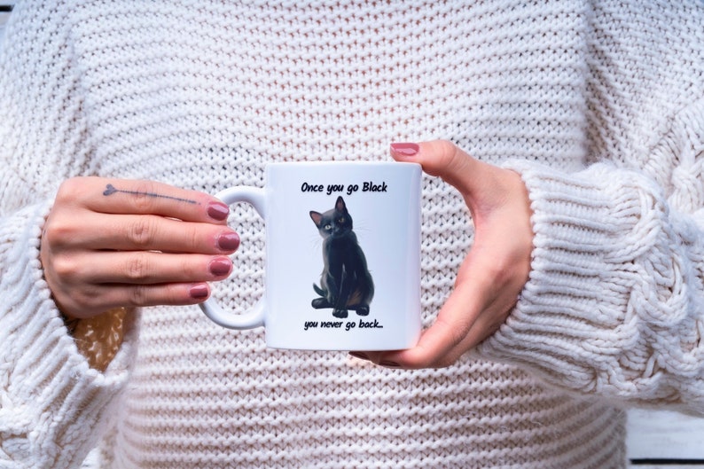 May include: White ceramic mug featuring a black cat illustration with the text "Once you go Black, you never go back...". The mug has a curved handle and is held by a person wearing a white sweater.