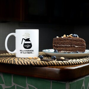 May include: White coffee mug with black text that reads "coffee" and "Hello darkness my old friend." The mug is sitting on a woven placemat next to a slice of chocolate cake with blueberries and nuts on top.
