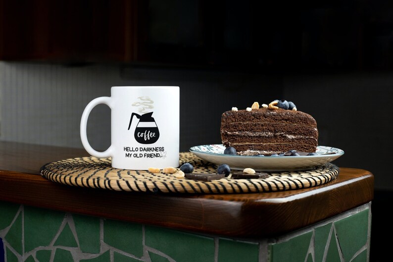 May include: White coffee mug with black text that reads "coffee" and "Hello darkness my old friend." The mug is sitting on a woven placemat next to a slice of chocolate cake with blueberries and nuts on top.