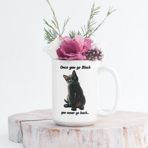 May include: White ceramic mug featuring a black cat illustration with the text "Once you go Black you never go back...". The mug is filled with a bouquet of pink, white, and purple flowers and greenery. The mug is sitting on a wooden surface.