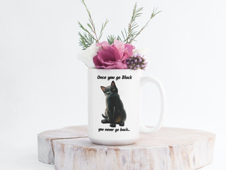 May include: White ceramic mug featuring a black cat illustration with the text "Once you go Black you never go back...". The mug is filled with a bouquet of pink, white, and purple flowers and greenery. The mug is sitting on a wooden surface.