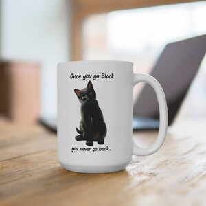 May include: White ceramic mug featuring a black cat illustration with the text "Once you go Black you never go back...". The mug has a large handle and is sitting on a wooden surface.