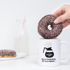 May include: White coffee mug with black text that reads "Hello darkness my old friend." and a black and white illustration of a coffee pot with steam coming out of it. The text "coffee" is written on the coffee pot. A hand is holding a chocolate donut with pink and yellow sprinkles over the mug.