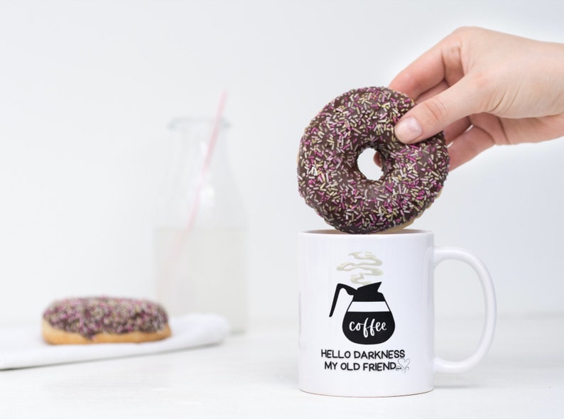 May include: White coffee mug with black text that reads "Hello darkness my old friend." and a black and white illustration of a coffee pot with steam coming out of it. The text "coffee" is written on the coffee pot. A hand is holding a chocolate donut with pink and yellow sprinkles over the mug.