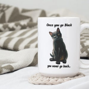 May include: White ceramic mug featuring a black cat illustration with the text "Once you go Black you never go back...". The mug has a large handle and is sitting on a beige coaster. The background includes a patterned blanket.