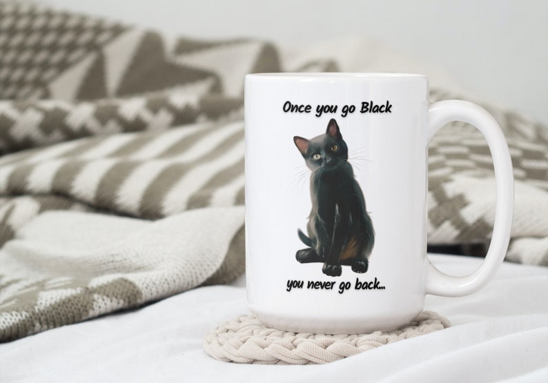 May include: White ceramic mug featuring a black cat illustration with the text "Once you go Black you never go back...". The mug has a large handle and is sitting on a beige coaster. The background includes a patterned blanket.