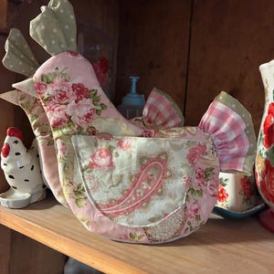 Op de afbeelding: Een stoffen theemuts in de vorm van een kip met een roze bloemenpatroon met rozen en een zakje met paisley-print. Het ontwerp bevat lichtgroene stippen op de kam en staart, samen met roze en wit gingham-stof op de staartveren.