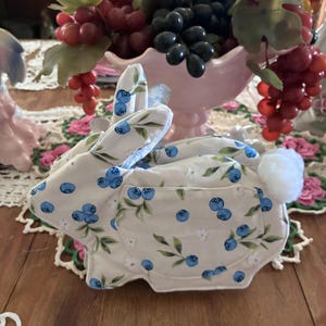 May include: A fabric rabbit figurine featuring a light beige background with a repeating pattern of blue blueberries and green leaves. The rabbit has a soft white pom-pom tail and a stitched pocket on its side, crafted from cotton-like material.