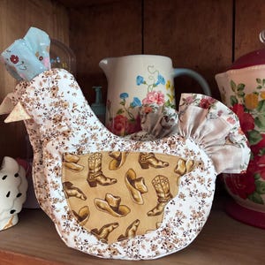 May include: A fabric chicken-shaped kitchen accessory featuring a white base with a brown floral print. The side panel displays a tan fabric with a repeating pattern of brown cowboy boots and hats. The piece includes a ruffled tail and a light blue comb.