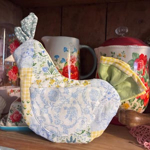 Puede incluir: Un artículo de cocina hecho a mano con forma de gallina, con telas de patchwork en estampados florales azules, cuadros amarillos y verdes. El artículo se exhibe en un estante de madera, con otros utensilios de cocina en el fondo.