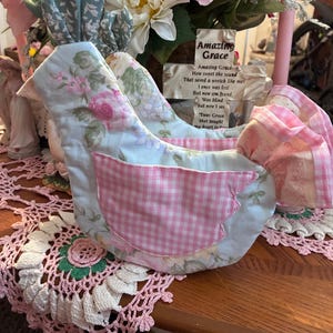 May include: A fabric bird ornament with a light blue body, pink gingham wings, and floral print details. The bird is placed on a pink lace doily. A sign with the words "Amazing Grace" is visible.