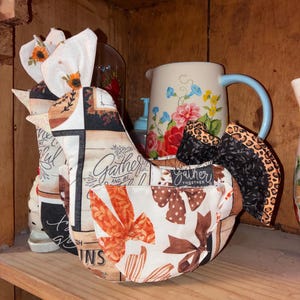May include: A fabric chicken-shaped ornament featuring a patchwork design with sunflowers, brown bows, and the text "Gather and be together." The chicken has a white and orange patterned pocket, a black and leopard print tail, and a white sunflower-print comb.