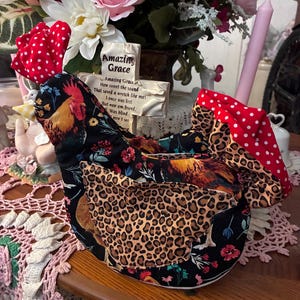 Könnte beinhalten: Ein hühnerförmiges Küchenaccessoire aus Stoff mit schwarzem Hintergrund, buntem Hahn- und Blumenmuster. Es verfügt über Stoffakzente mit Leopardenmuster an den Flügeln sowie einen Kamm und einen Schwanz aus rot-weiß gepunktetem Stoff.