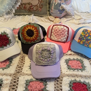 May include: Five colorful trucker hats with crocheted square designs. The hats feature a variety of colors, including white, black, pink, blue, and lavender. The crocheted squares display floral and geometric patterns.