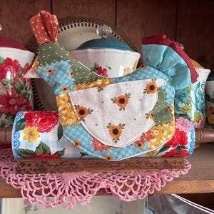 Pode incluir: Acessório de cozinha artesanal em forma de galinha, provavelmente uma capa para rolo de massa, feito de tecido patchwork em azul, verde e laranja. A galinha tem um avental branco com detalhes de girassóis. Um rolo de massa com tecido floral é visível.