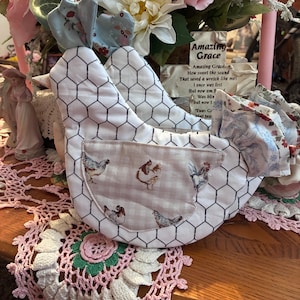 Pode incluir: Um ornamento de tecido em forma de galinha com um padrão de rede de galinheiro preta sobre fundo branco. O corpo inclui um bolso bege com uma estampa de galinhas, enquanto a cabeça e a cauda são acentuadas com tecido floral azul claro.