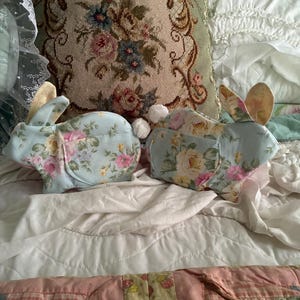 Pode incluir: Duas decorações de coelho de pelúcia com tecido estampado floral em tons de azul, rosa e amarelo. Os coelhos têm orelhas compridas e caudas brancas fofas. Eles estão em uma cama com uma almofada floral e um edredom de retalhos.