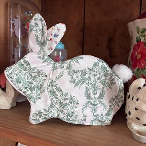 Pode incluir: Uma cesta de tecido em forma de coelho da Páscoa. O coelho é principalmente rosa claro com um padrão floral verde. As orelhas têm um padrão floral com uma cauda branca e fofa. A cesta está numa prateleira de madeira.
