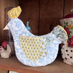 Peut inclure: Objet en forme de poulet en tissu, probablement pour la cuisine. Le corps est blanc avec un motif floral bleu, tandis que l'aile et la crête présentent des motifs floraux et vichy jaunes. Il repose sur une étagère en bois.