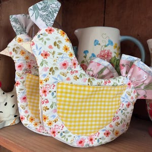 Puede incluir: Dos accesorios de cocina de tela con forma de gallina, con base blanca y un estampado floral colorido de flores rosas, amarillas y azules. Cada gallina tiene un bolsillo de tela de cuadros amarillos y blancos en el lateral y una cola con volantes rosa.
