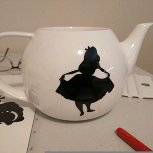Pu&ograve; includere: Teiera in ceramica bianca con una silhouette nera di una ragazza con un vestito. La teiera ha un corpo arrotondato, un manico curvo e un beccuccio. Il design &egrave; probabilmente ispirato a un personaggio di fiaba.
