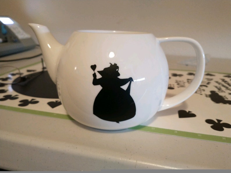 Pu&ograve; includere: Teiera in ceramica bianca con corpo arrotondato, manico curvo e beccuccio. Sul davanti c'&egrave; una silhouette nera di una regina che tiene un cuore. La teiera &egrave; su una superficie bianca.