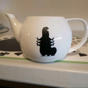 Pu&ograve; includere: Teiera in ceramica bianca con una silhouette nera di bruco. La teiera ha un manico curvo, un beccuccio e un corpo arrotondato. Il disegno del bruco &egrave; sul davanti. La teiera &egrave; su una superficie bianca.