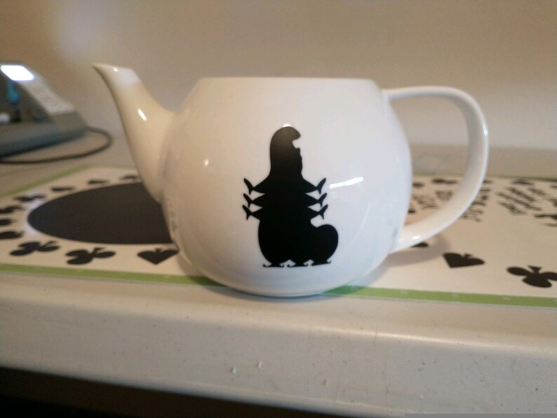Pu&ograve; includere: Teiera in ceramica bianca con una silhouette nera di bruco. La teiera ha un manico curvo, un beccuccio e un corpo arrotondato. Il disegno del bruco &egrave; sul davanti. La teiera &egrave; su una superficie bianca.