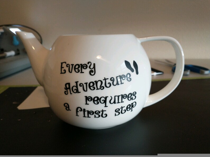 Pu&ograve; includere: Teiera in ceramica bianca con corpo arrotondato, beccuccio e manico. La teiera presenta un testo nero che recita "Every Adventure requires a first step" con impronte nere. Un affascinante accessorio da cucina.