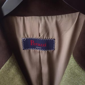 May include: Close-up of the interior of a green and brown blazer. The lining is beige, and a blue label with the text "Perazzi classic" is sewn into the jacket. The blazer is hanging on a black hanger.