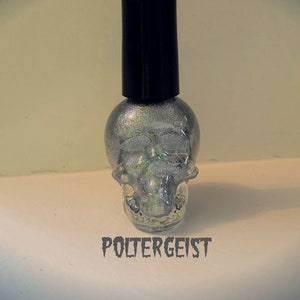 May include: A silver glitter nail polish bottle shaped like a skull with a black cap. The bottle is labeled "Poltergeist".