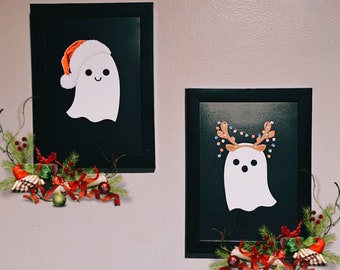 Può includere: Due immagini incorniciate nere con illustrazioni di fantasmi bianchi. Il fantasma a sinistra indossa un cappello di Babbo Natale rosso. Il fantasma a destra ha le corna di renna e una ghirlanda di luci. Entrambe le immagini sono decorate con decorazioni natalizie rosse e verdi.