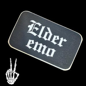 Könnte beinhalten: Ein schwarzes rechteckiges Schild mit den Worten "Elder Emo" in weißer gotischer Schrift. Eine weiße Skeletthand, die ein Peace-Zeichen macht, befindet sich in der linken unteren Ecke des Bildes.