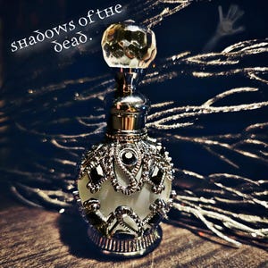 May include: A silver and clear glass bottle with a decorative design and a crystal stopper. The bottle is sitting on a wooden surface. The text "shadows of the dead." is visible in the background.