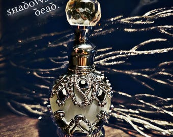 May include: A silver and clear glass bottle with a decorative design and a crystal stopper. The bottle is sitting on a wooden surface. The text "shadows of the dead." is visible in the background.