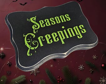 Puede incluir: Un letrero de madera negro con un borde blanco y una inscripción verde que dice "Seasons Creepings". El letrero está rodeado de ramas de pino verdes, bayas rojas y copos de nieve plateados sobre un fondo burdeos.
