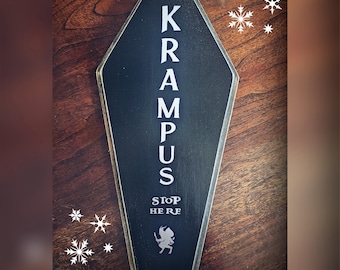 Puede incluir: Un letrero de madera negro en forma de ataúd con la palabra "KRAMPUS" en letras blancas. Debajo, se lee "STOP HERE" con una pequeña silueta de una figura de Krampus. Copos de nieve decorativos en el fondo.
