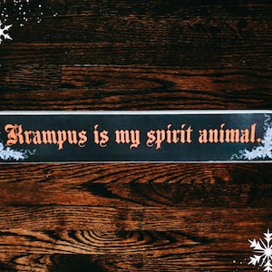 May include: A black sign with white floral accents and the text "Krampus is my spirit animal." The sign is on a dark wood background.