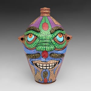 May include: A ceramic jug shaped like a face with a purple, green, and tan patterned surface. It features raised eyebrows, a mustache, and a goatee, with orange eyes and a wide, toothy grin. The vessel has a narrow neck and two side handles.
