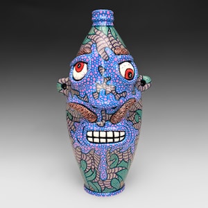 May include: A ceramic bottle shaped like a face with a blue base, pink polka dots, and intricate black line patterns. The face features two large red eyes, a textured brown mustache, and a wide mouth with visible white teeth and small ears.