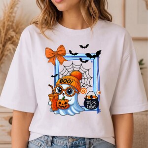 May include: A white t-shirt featuring a graphic of a ghost wearing an orange beanie with the text BOO!, holding a pumpkin and a drink. The design includes a spiderweb, bats, an orange bow, and a black cauldron with the text trick or treat.