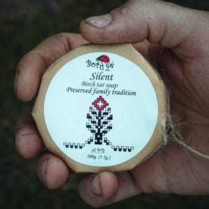 Può includere: Un sapone rotondo, marrone, avvolto in carta con un'etichetta bianca. L'etichetta presenta un logo di coccinella rossa, il testo "Borde Silent", e le parole "Sapone di catrame di betulla Tradizione familiare preservata". L'etichetta presenta anche un design stilizzato di albero in bianco e nero e il testo "APB 100g. (±5g.)".